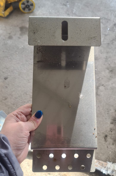 Szlifowany element metalowy z otworami, trzymany w dłoni. Widoczny fragment wózka warsztatowego w tle. Detal konstrukcyjny z blachy nierdzewnej.