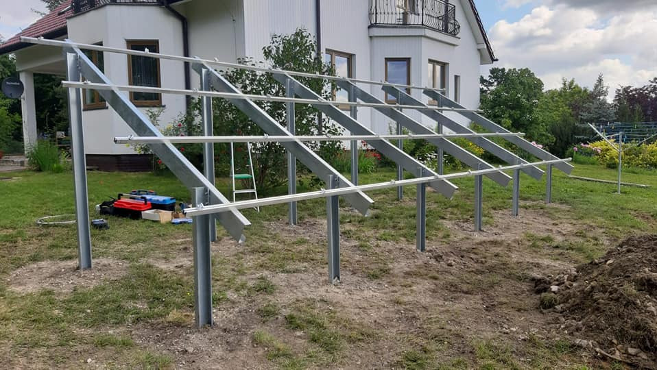 Stalowa konstrukcja pergoli w ogrodzie przed domem. Widoczne słupy i poprzeczki. Na trawie leżą narzędzia w skrzynkach. Częściowo zniwelowany teren.