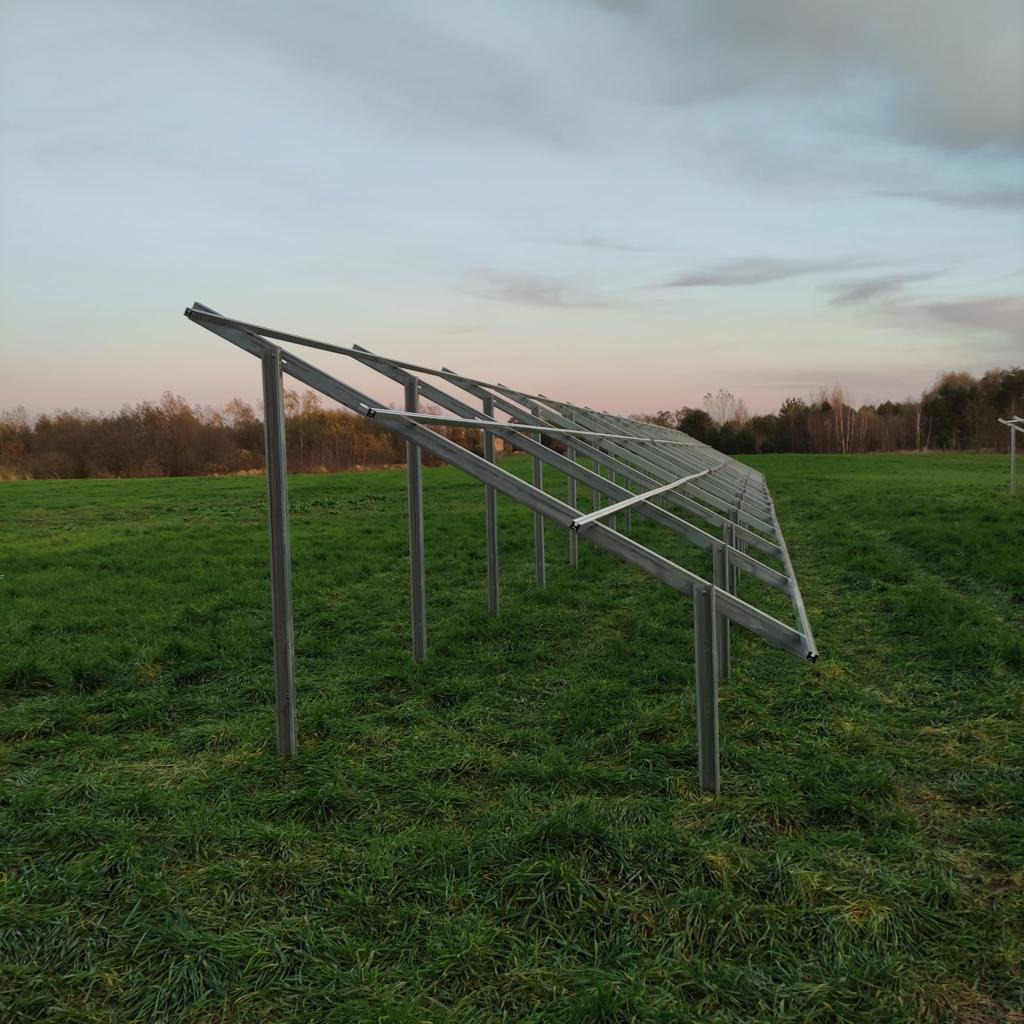 Metalowa konstrukcja wsporcza na łące, prawdopodobnie pod panele fotowoltaiczne. Widoczne elementy montażowe i naturalne otoczenie.