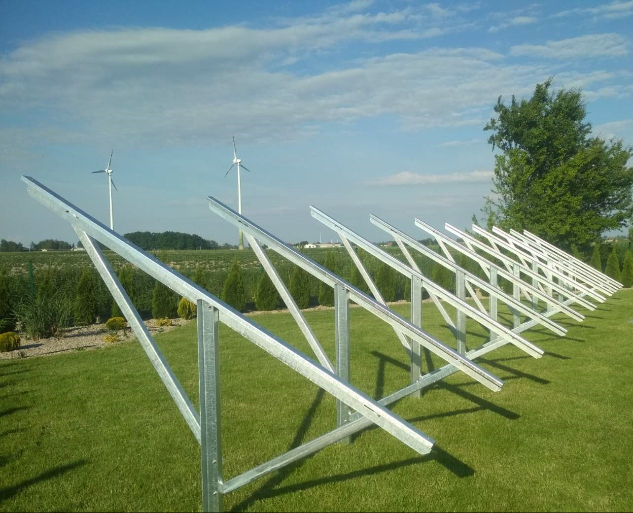 Metalowa konstrukcja wsporcza na trawniku w Złotowie, z turbinami wiatrowymi w tle. Solidne, spawane elementy, regularny układ, jasne niebo.