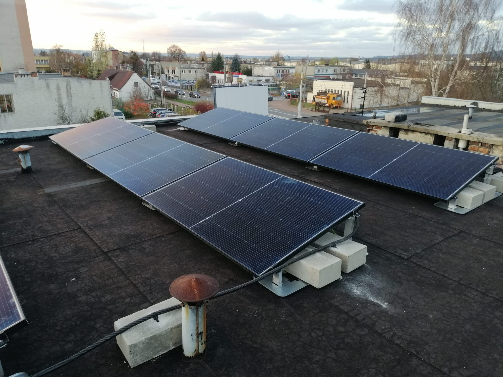 Panele słoneczne zamontowane na płaskim dachu budynku, widoczne betonowe podpory i przewody. W tle zabudowa miejska i pochmurne niebo.