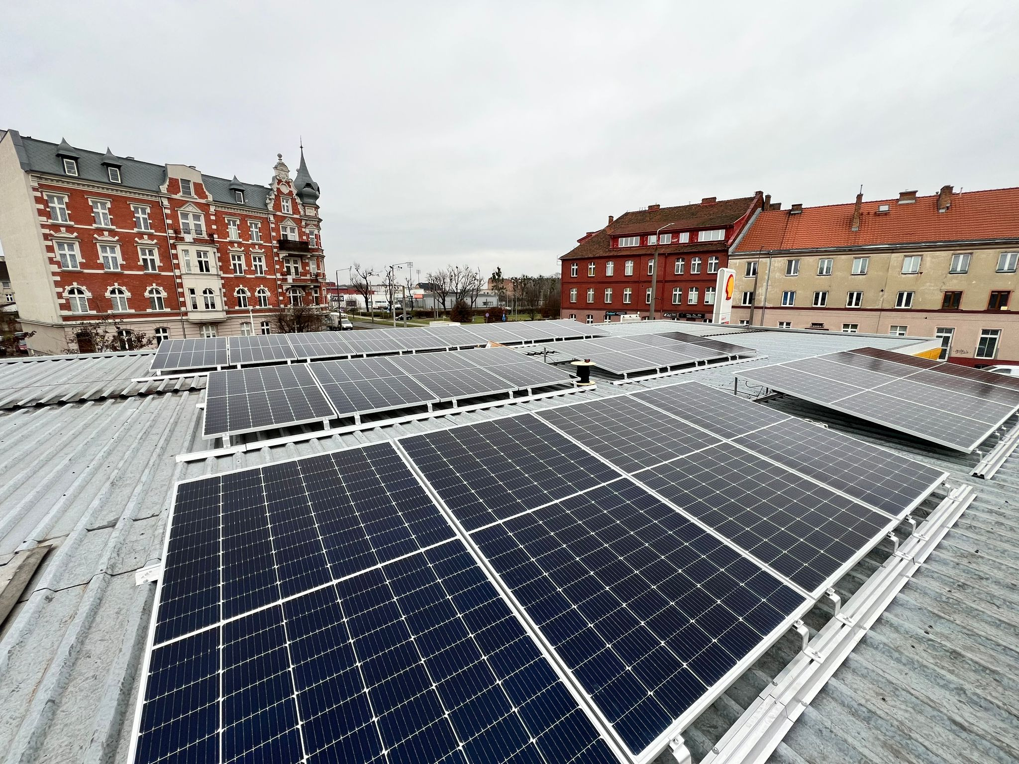 Rząd paneli słonecznych na dachu budynku w pochmurny dzień. W tle widoczne budynki miejskie z czerwonej cegły i logo stacji paliw.