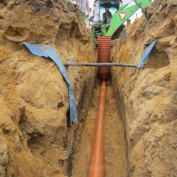 SKYWEATHER SPÓŁKA Z OGRANICZONĄ ODPOWIEDZIALNOŚCIĄ - Pomarańczowa rura w wykopie, zabezpieczona niebieską folią. Na górze koparka i dwie osoby nadzorujące prace. Widok z góry na instalację kanalizacyjną.