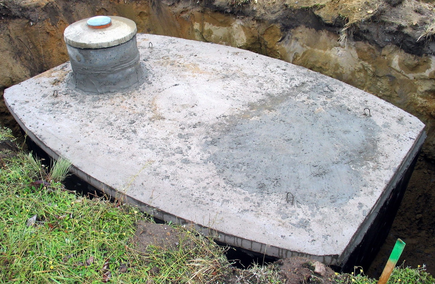 Betonowy szambo częściowo wkopany w ziemię, widoczny właz z pokrywą, trawa i ziemia wokół. Ujęcie z bliska, ukazujące detale konstrukcji.