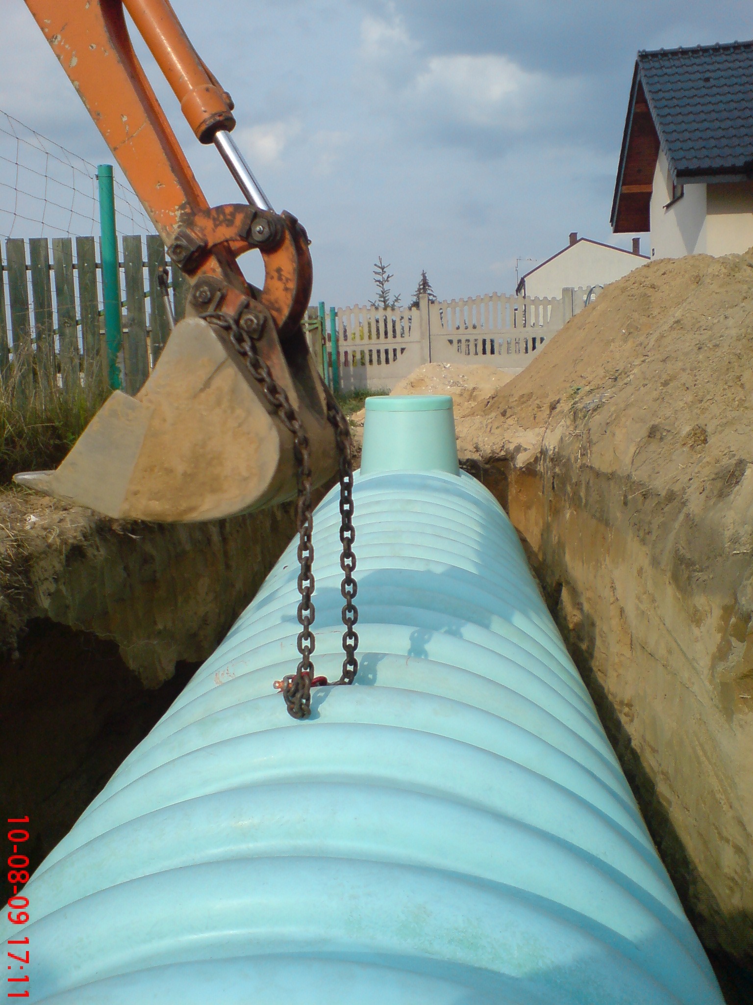 Montaż jasnoniebieskiego szamba z tworzywa sztucznego w wykopie, z koparką w tle. Widoczny fragment zabudowy jednorodzinnej i ogrodzenia.