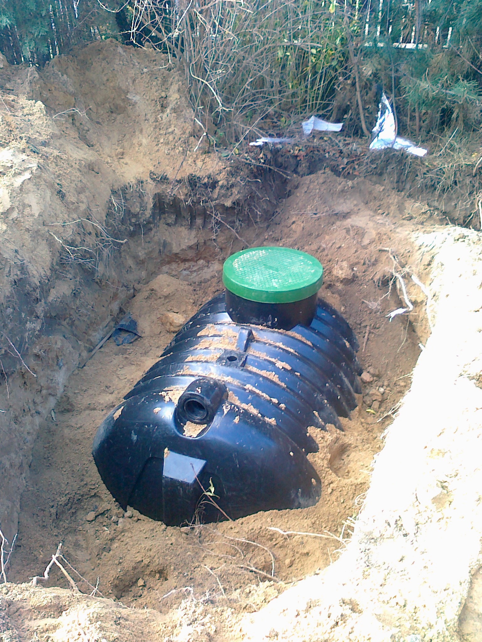 Czarny zbiornik z zieloną pokrywą w wykopie ziemnym. Widoczne ślady ziemi na zbiorniku i ścianach wykopu. Tło stanowią drzewa i krzewy.