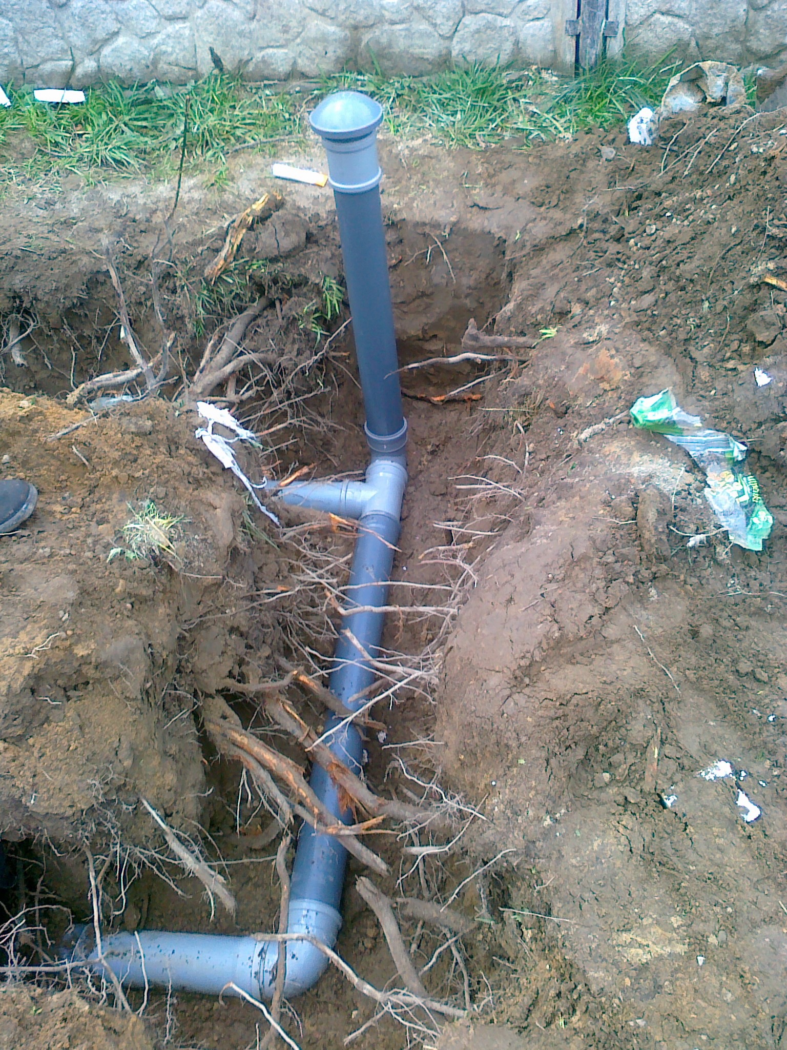 Szare rury kanalizacyjne ułożone w wykopie ziemnym, widoczne korzenie drzew i fragment muru w tle. Instalacja w trakcie montażu.