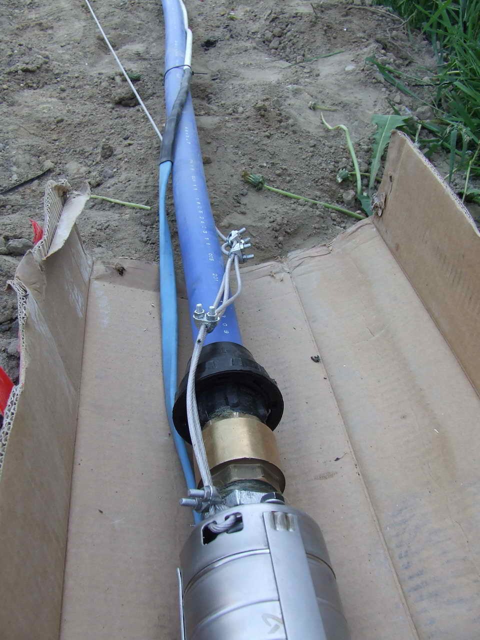 Niebieska rura z kablem i metalową pompą w kartonowym wykopie, prawdopodobnie element instalacji wodnej lub kanalizacyjnej na etapie montażu.