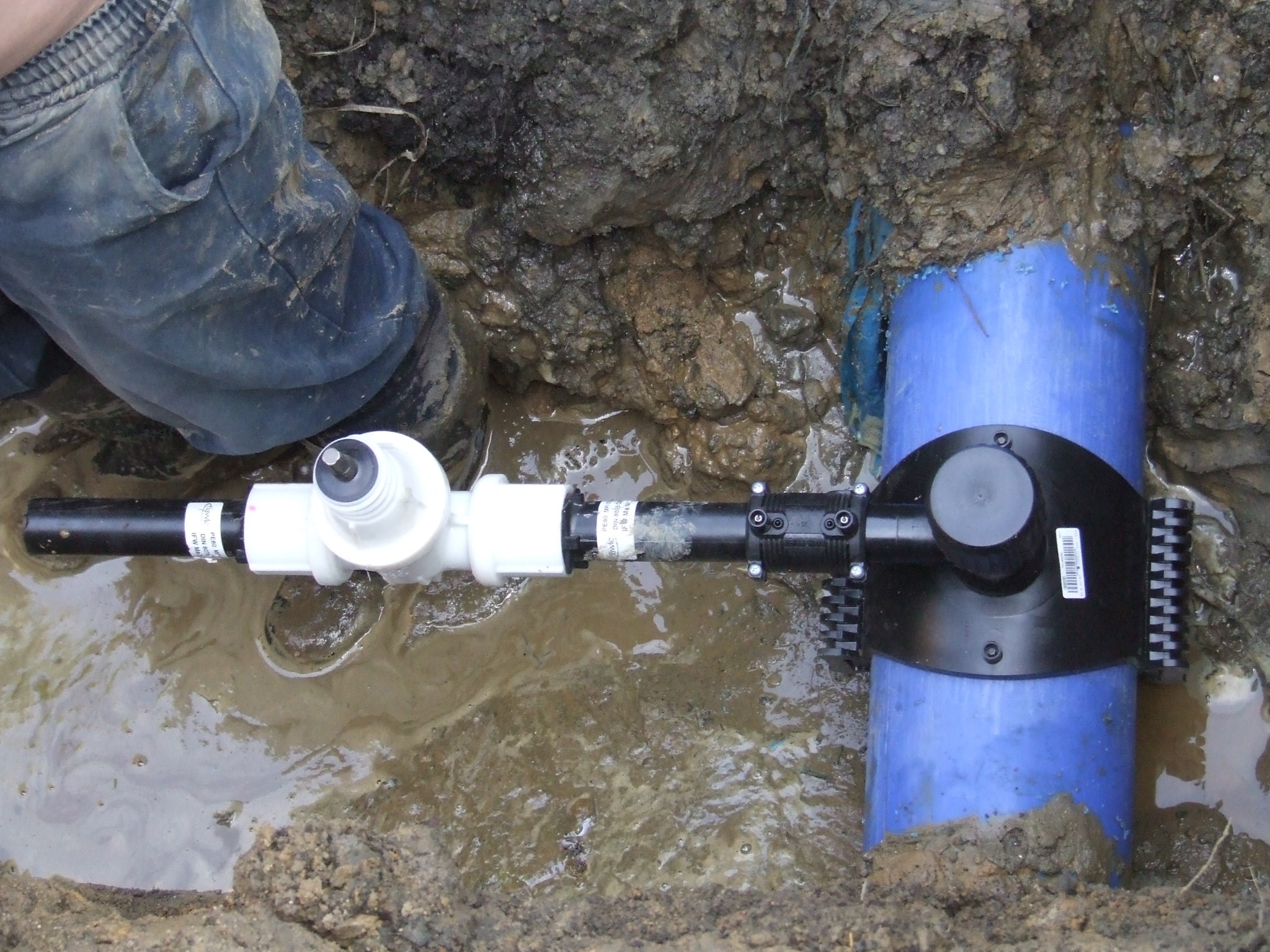 Podłączenie rury wodociągowej do niebieskiej rury w wykopie ziemnym wypełnionym wodą. Widoczne elementy złączek i instalacji. Częściowo widoczna noga osoby w roboczych spodniach.
