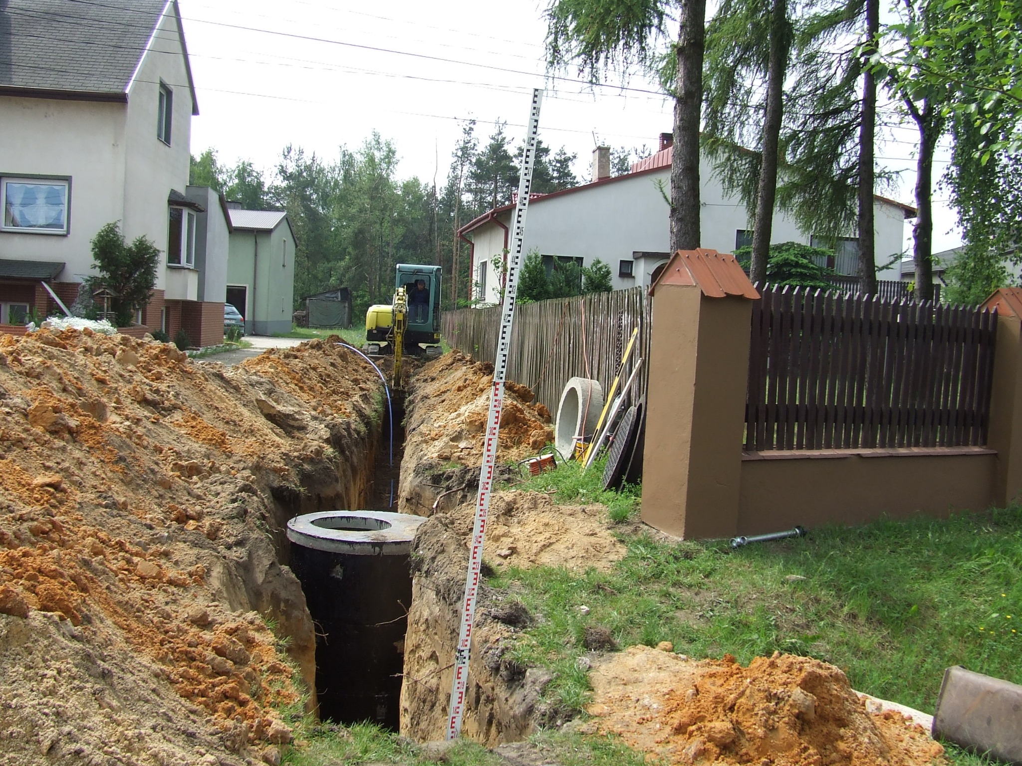 Montaż betonowego szamba w wykopie, widoczny mini-koparka, poziomica i elementy konstrukcyjne. Prace ziemne w toku, w tle domy jednorodzinne.