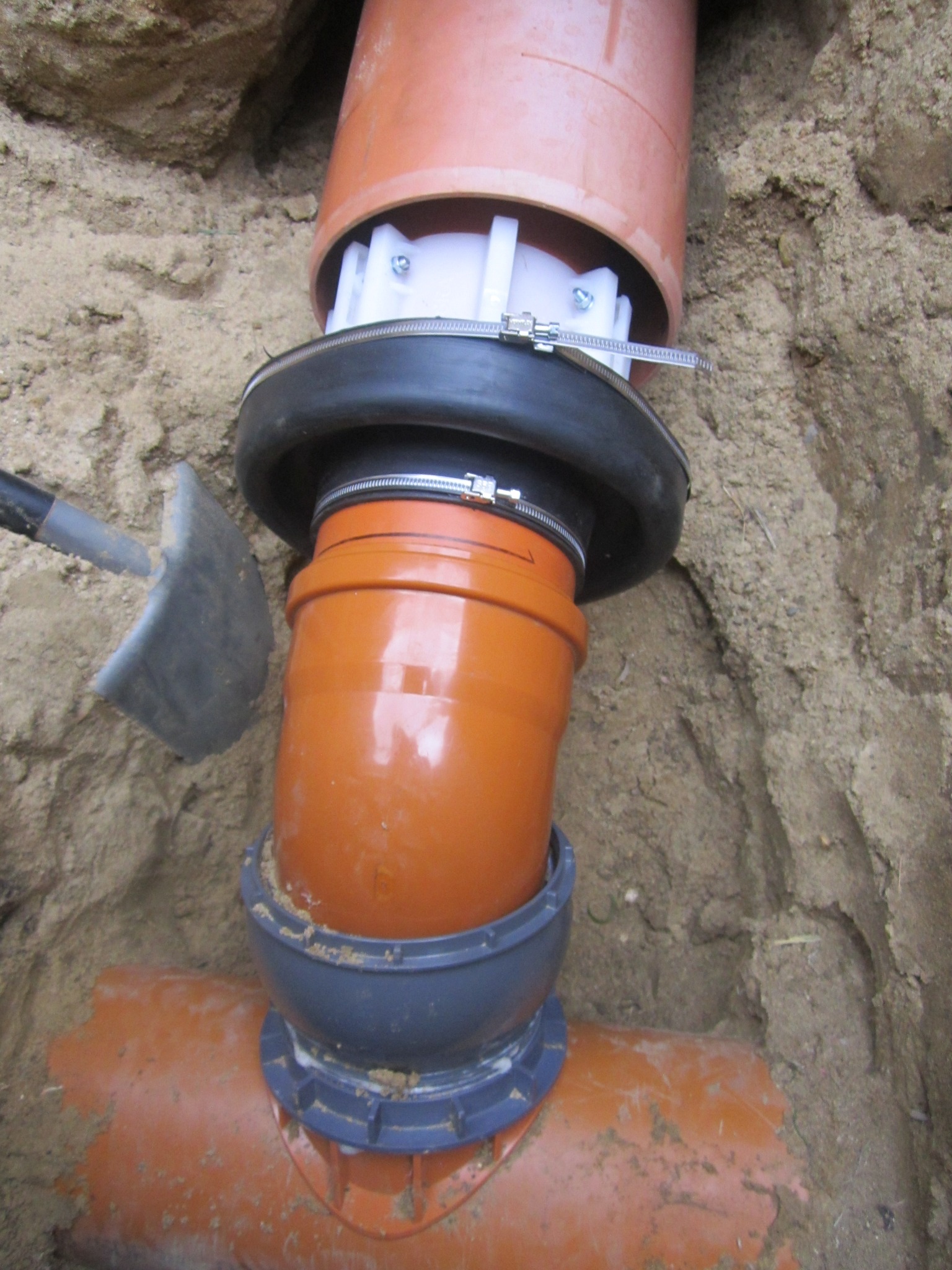 Pomarańczowe rury kanalizacyjne łączone w wykopie, widoczne złącza, obejmy i fragment łopaty. Realizacja przyłącza wod-kan.