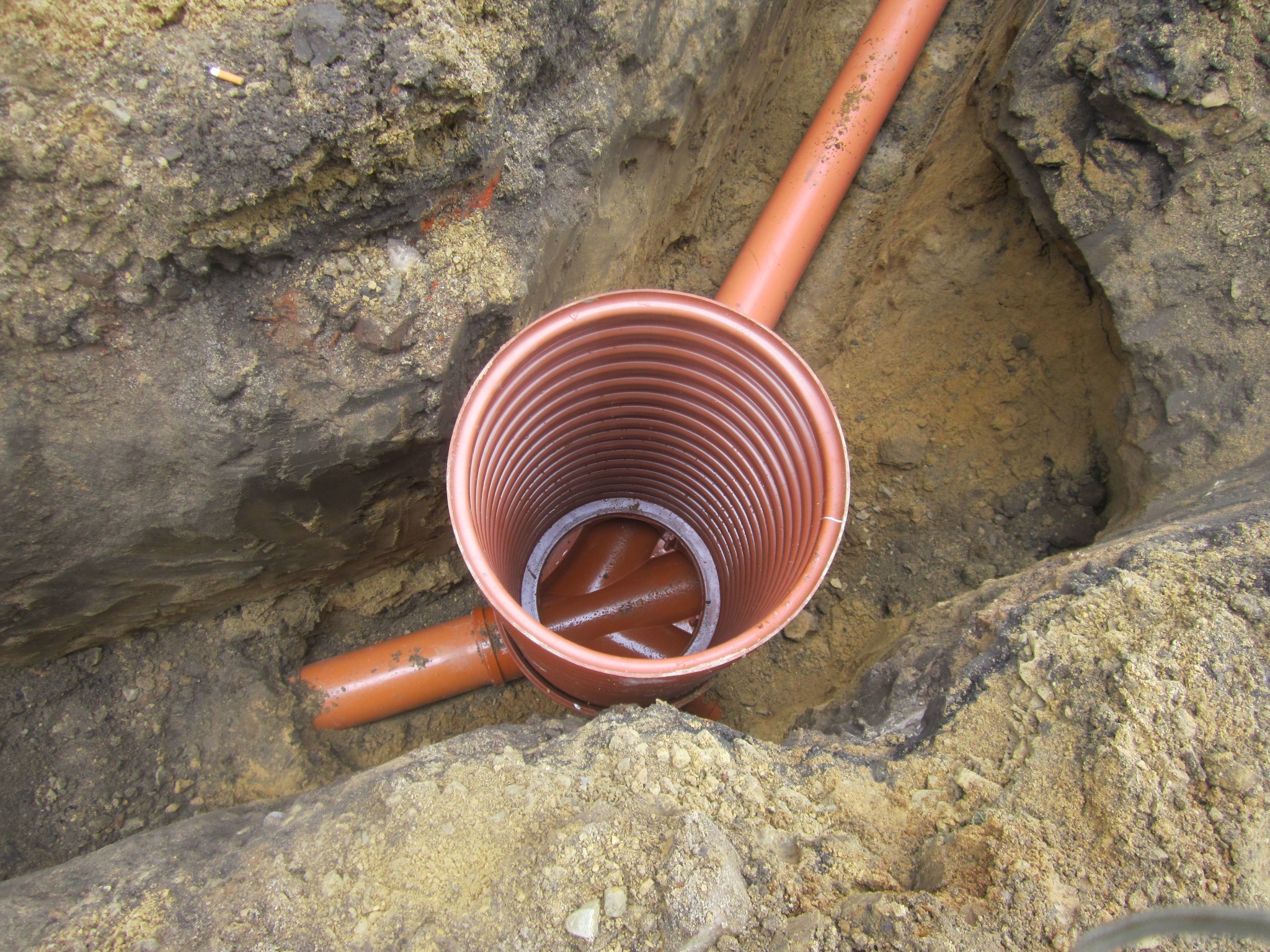 Pomarańczowa rura kanalizacyjna w wykopie z widocznym włazem rewizyjnym. Ziemia wokół wykopu o zróżnicowanej strukturze i kolorystyce. Widoczne połączenia rur.