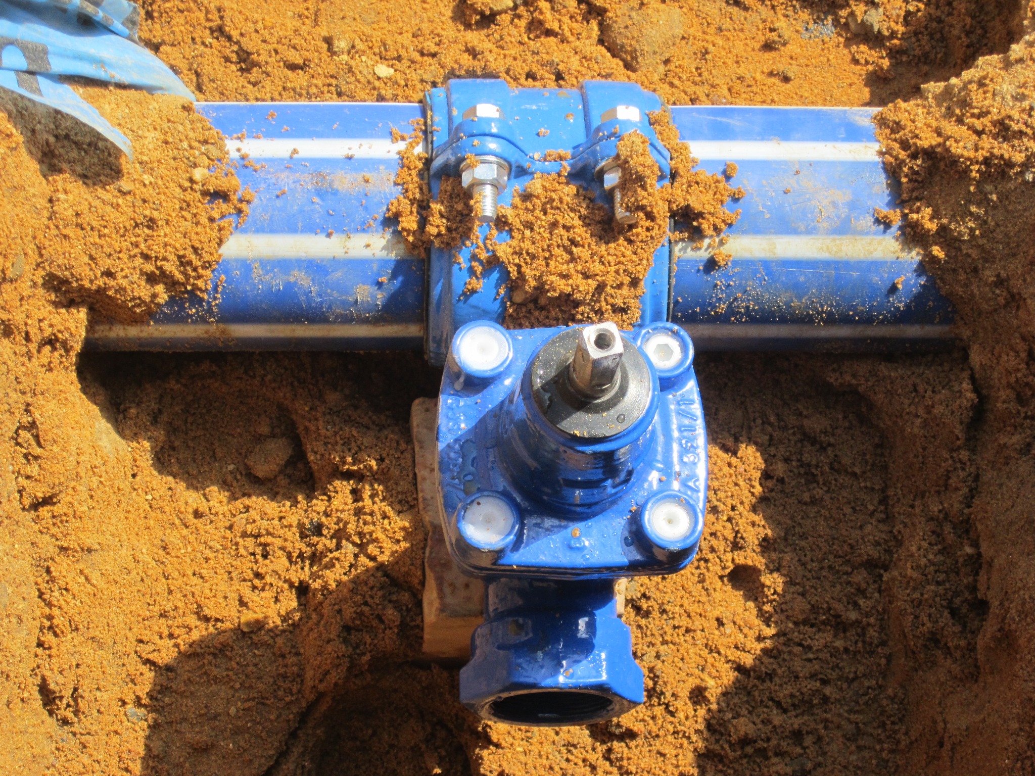 Niebieski zawór na rurze wodociągowej w wykopie, otoczony piaskiem. Widoczne śruby mocujące i fragment rury z niebieskimi pasami.