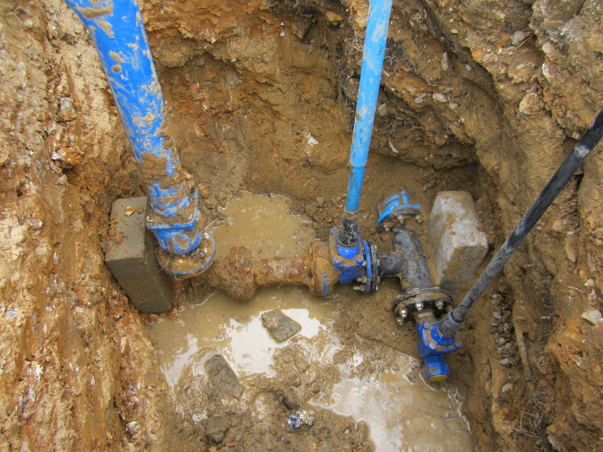 Niebieskie rury i zawory w wykopie wypełnionym wodą i błotem. Fragment instalacji wodociągowej z widocznymi elementami złącznymi i podparciem z betonowych bloczków.