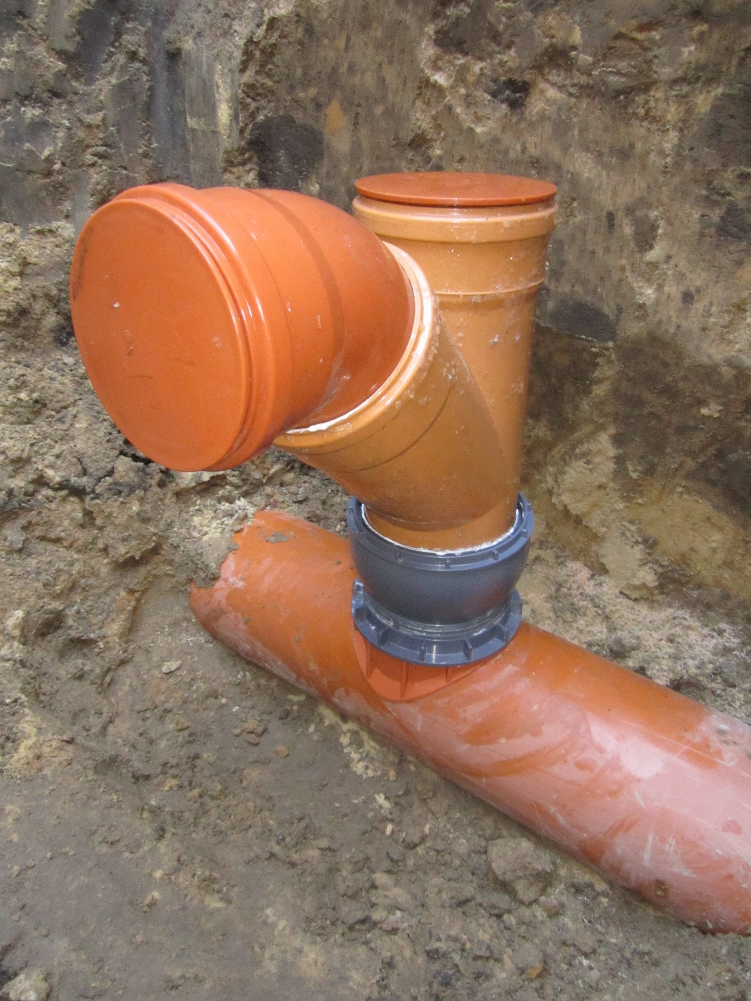 Pomarańczowe rury kanalizacyjne z łącznikiem w ziemi, widoczne w wykopie. Element instalacji sanitarnej.