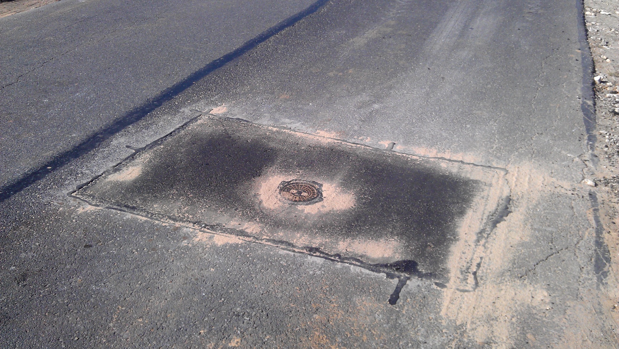 Studzienka kanalizacyjna w asfalcie, otoczona piaskiem. Widoczny fragment drogi i pobocza. Kątowy kadr podkreśla fakturę nawierzchni.