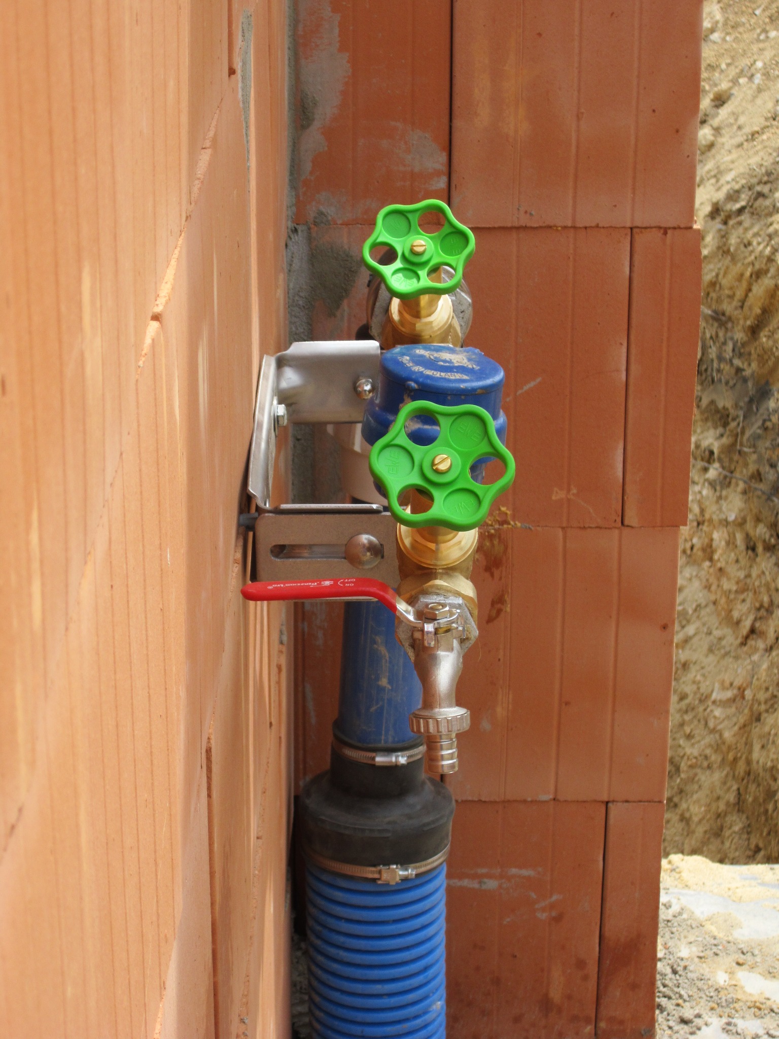 Instalacja wodna z zaworami i niebieskim karbowanym wężem, zamocowana do ściany z cegły dziurawki w Żorach. Widoczne elementy montażowe i przyłącza.