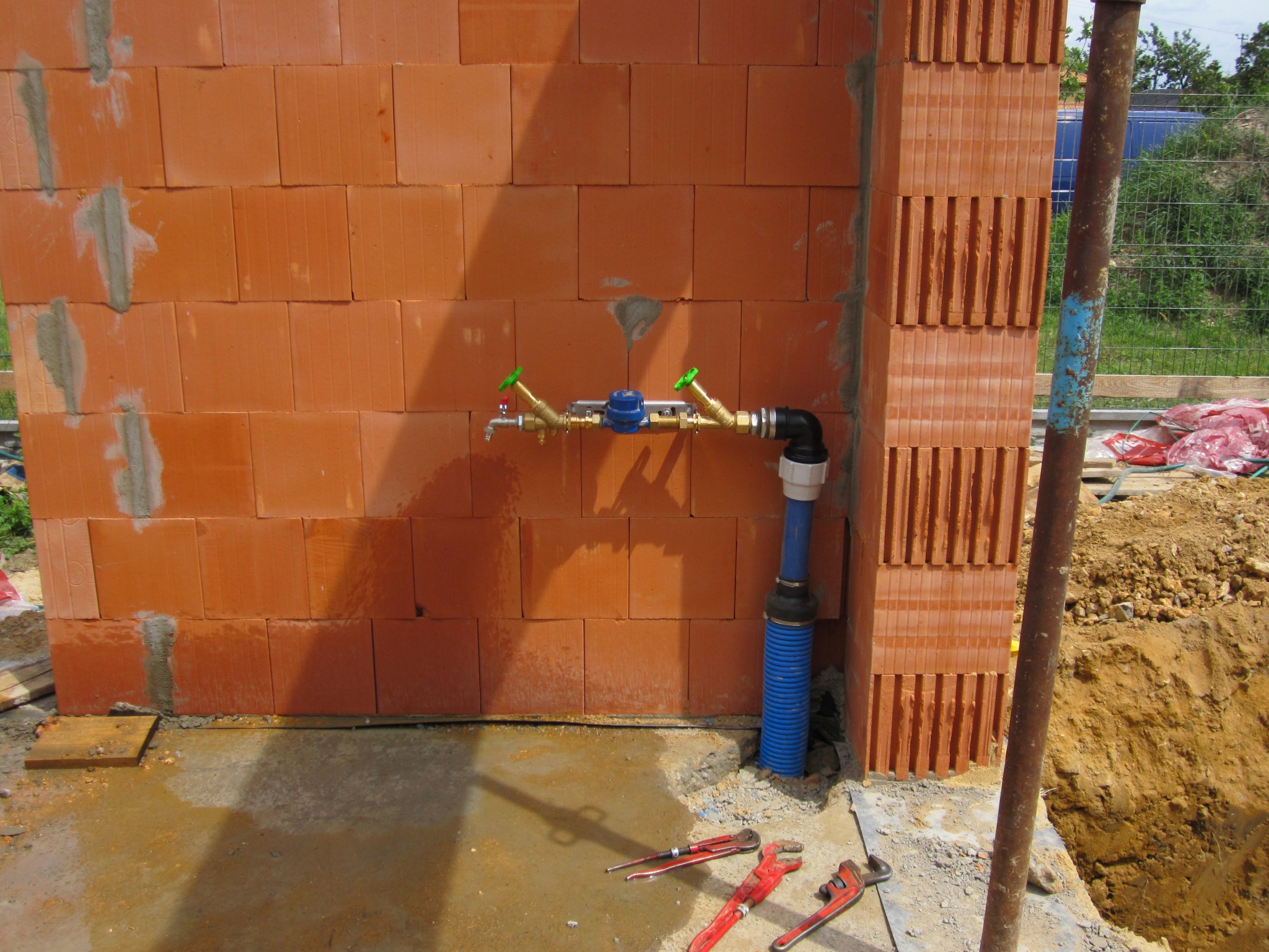 Instalacja wodna z zaworami i licznikiem, zamontowana na ścianie z czerwonej cegły. Na podłodze leżą narzędzia hydrauliczne, obok wykop pod fundament.