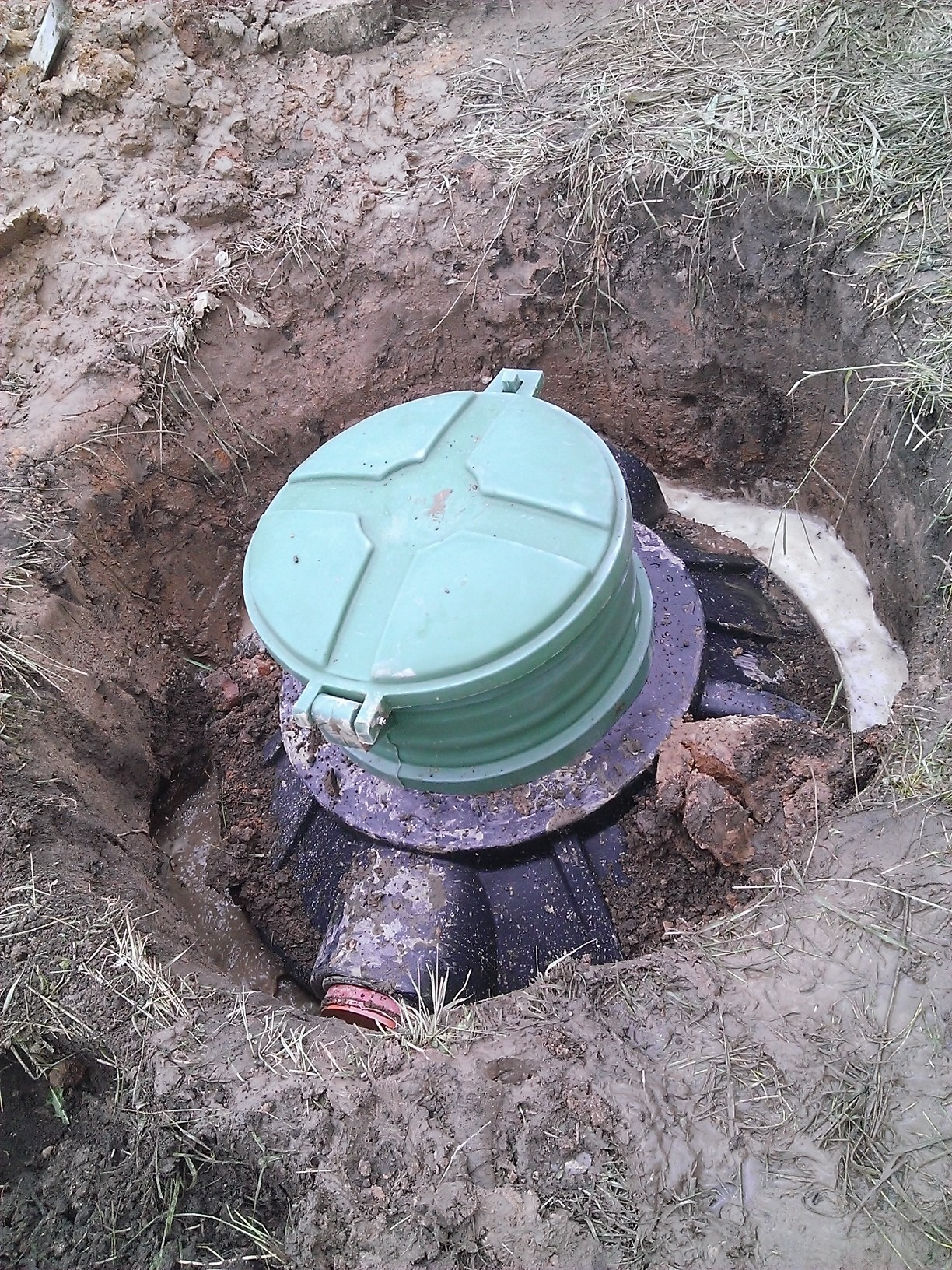 Zielona pokrywa studzienki kanalizacyjnej częściowo zasypana ziemią w wykopie. Widoczne ślady wody. Praca w toku.