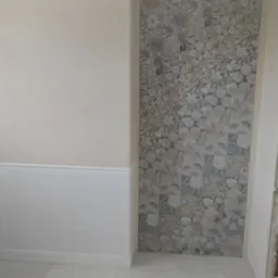 Jasna łazienka z dekoracyjną ścianą z płytek w kwiatowe wzory, białą toaletą i fragmentem ościeżnicy drzwi. Widoczne wykończenie w stylu klasycznym.