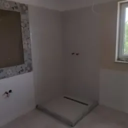 Wnętrze łazienki w trakcie prac: przygotowana ściana pod płytki, zamontowany odpływ liniowy w brodziku. Widoczne przyłącza wodne i częściowo zamontowana toaleta.