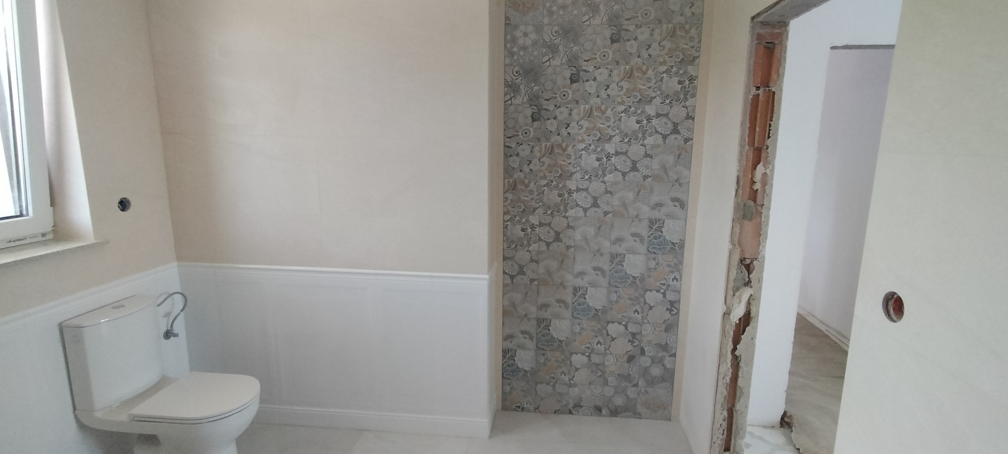 Jasna łazienka z dekoracyjną ścianą z płytek w kwiatowe wzory, białą toaletą i fragmentem ościeżnicy drzwi. Widoczne wykończenie w stylu klasycznym.