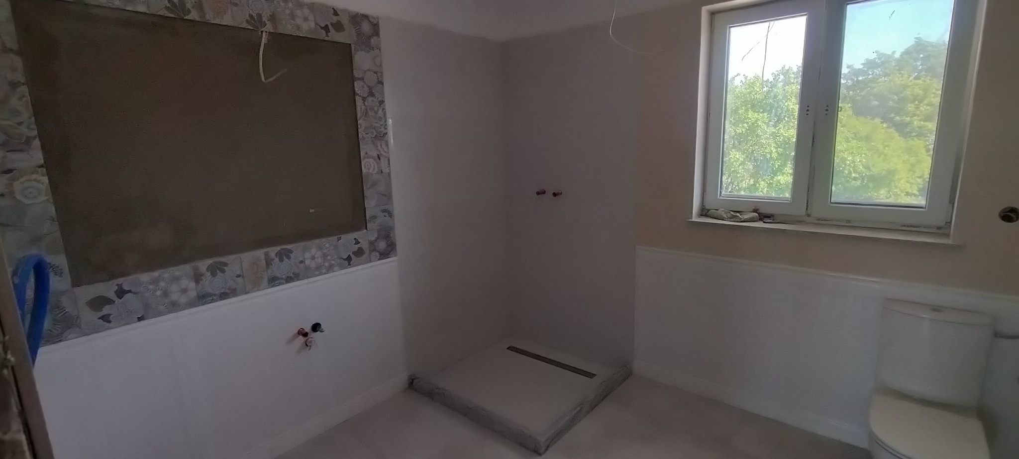 Wnętrze łazienki w trakcie prac: przygotowana ściana pod płytki, zamontowany odpływ liniowy w brodziku. Widoczne przyłącza wodne i częściowo zamontowana toaleta.