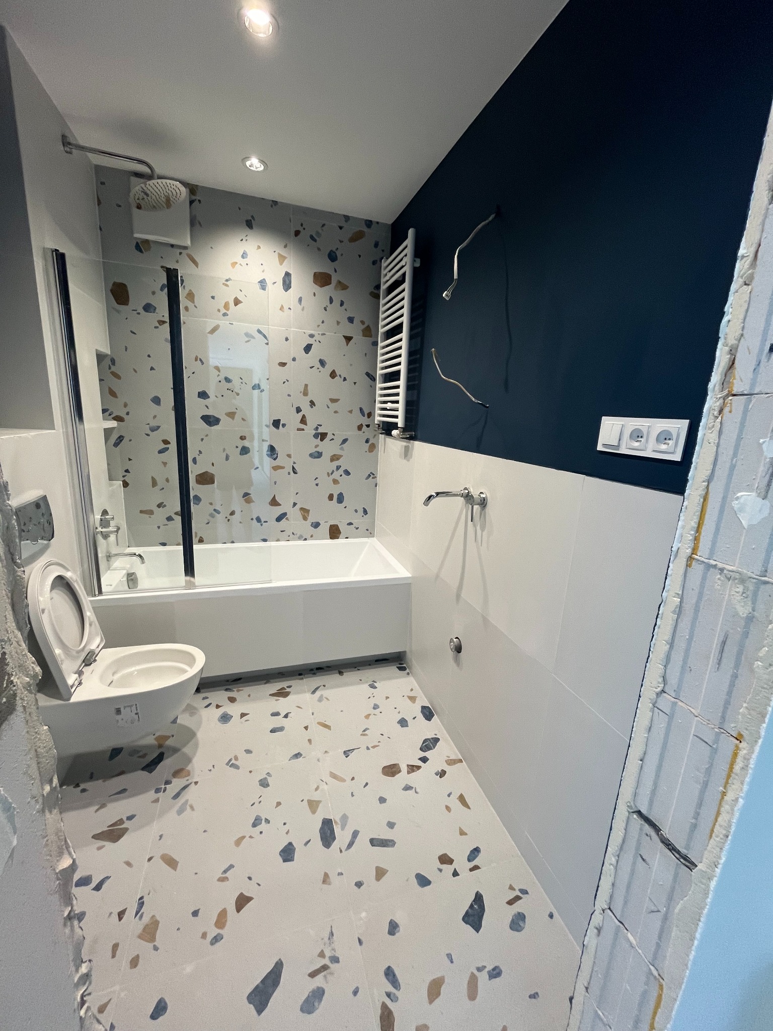 Nowoczesna łazienka po remoncie z wanną za szklanym parawanem, toaletą i grzejnikiem drabinkowym na tle ściany w kolorze granatowym i białych płytek.