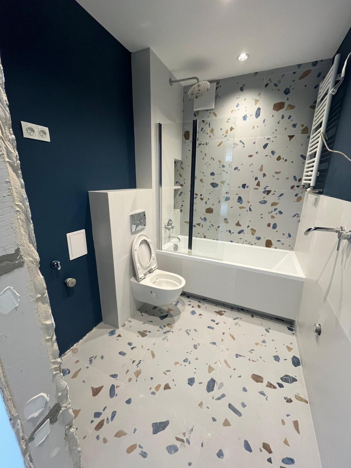 Wykończona łazienka z wanną za szybą, wiszącą toaletą i wzorzystymi płytkami na podłodze i ścianie. Ściana pomalowana na granatowo, widoczne gniazdka.