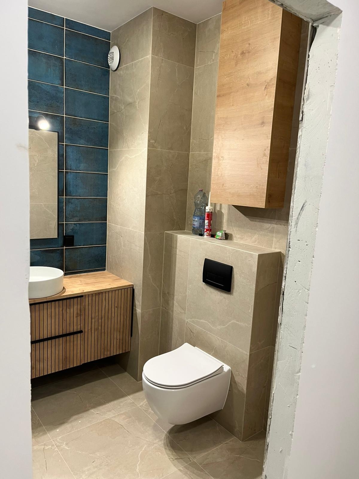 Wykończona łazienka z wiszącą toaletą, umywalką na drewnianej szafce i niebieskimi płytkami na ścianie. Widoczna armatura i akcesoria łazienkowe.
