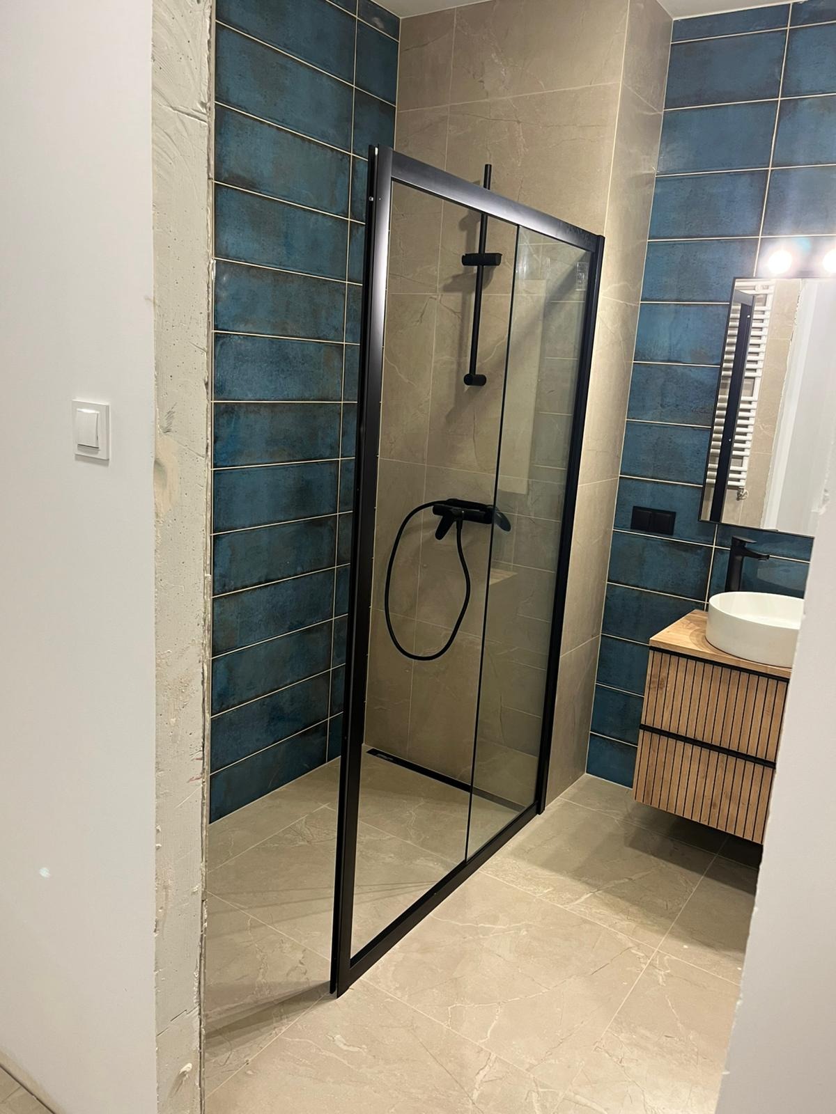 Wykończona łazienka z kabiną prysznicową typu walk-in z czarną ramą, umywalką nablatową na szafce z lamelami i niebieską glazurą na ścianach.