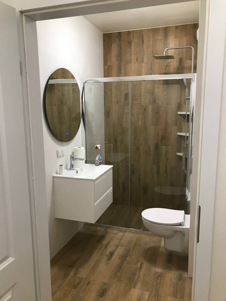 Nowoczesna łazienka z drewnopodobnymi płytkami, owalnym lustrem z podświetleniem LED i kabiną prysznicową ze szklanymi drzwiami przesuwnymi.