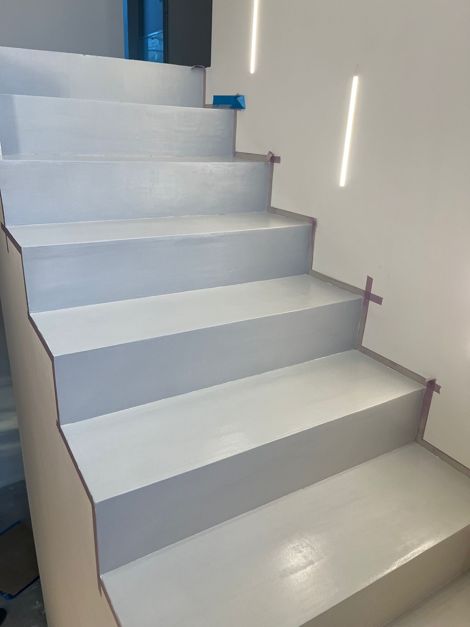 Świeżo pomalowane, jasnoszare schody wewnętrzne z taśmą malarską zabezpieczającą krawędzie. Minimalistyczne oświetlenie w tle.