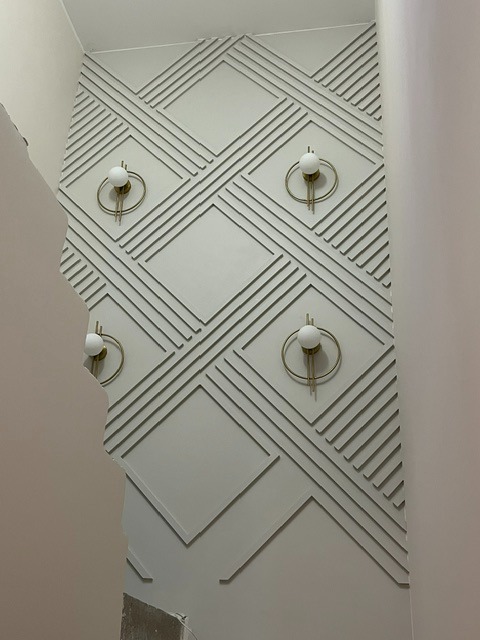 Ściana z dekoracyjnymi listwami w geometryczny wzór rombów i pasków. Na ścianie zamontowane dwa złote kinkiety z okrągłymi, białymi kloszami. Wykończenie wnętrz.