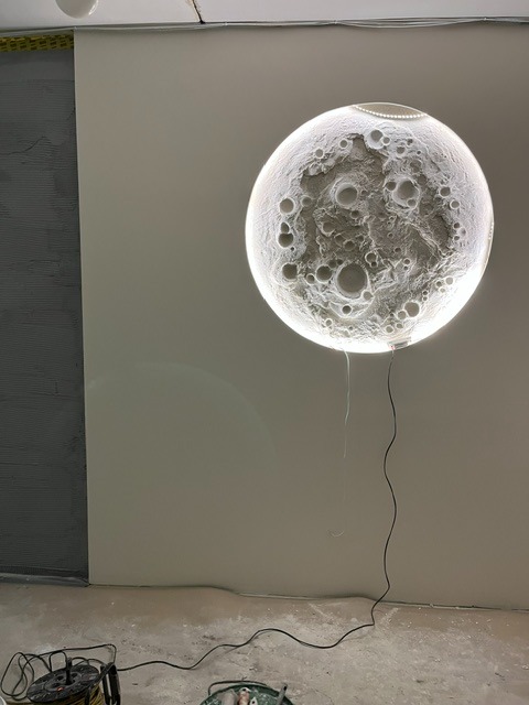Nietypowa lampa ścienna w kształcie księżyca z wyraźną fakturą kraterów, zawieszona na świeżo pomalowanej ścianie. Widoczne narzędzia malarskie i kabel zasilający.