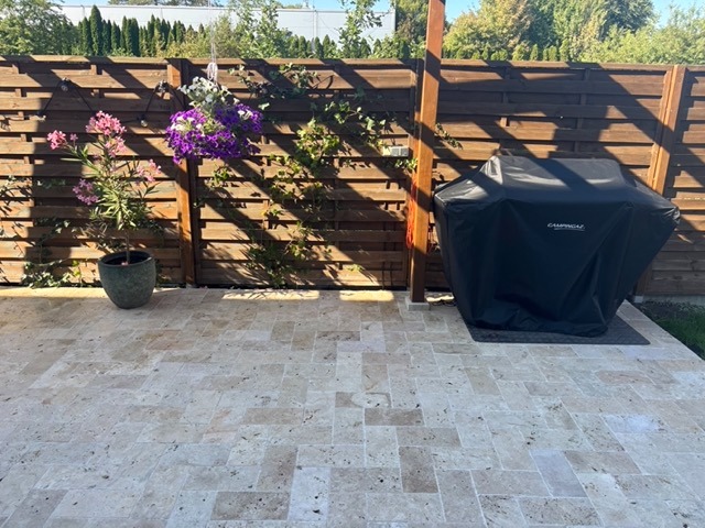 Jasny trawertyn na patio z grillem CampinGaz pod drewnianą pergolą, kwiaty w donicach i wiszących koszach. Słoneczny dzień.