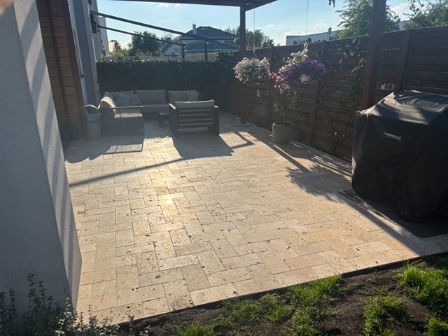 Ułożona jasna kostka na patio z meblami ogrodowymi, zielenią i grillem. Widok na dom z pergolą i wiszącymi kwiatami w słoneczny dzień.