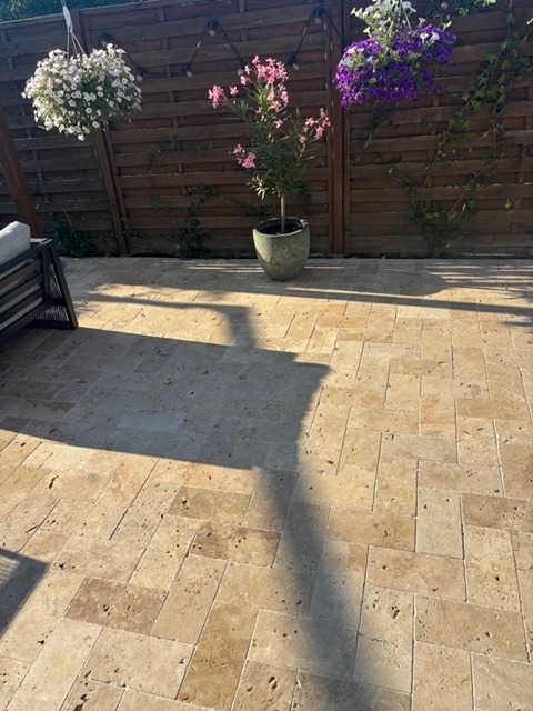 Ułożona trawertynowa podłoga na tarasie w Warszawie. Widoczny wzór jodełki, drewniane ogrodzenie z kwiatami i cień. Ciepłe, popołudniowe światło.