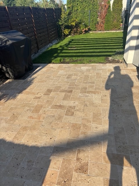 Ułożona jasna kostka brukowa na tarasie, widoczny cień osoby. W tle trawnik, drewniany płot i roślinność.