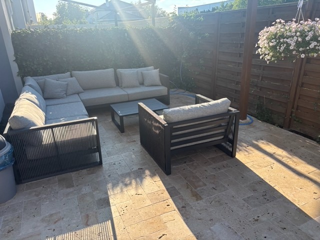 Umeblowany taras z narożną sofą, fotelem i stolikiem kawowym na beżowej, kamiennej podłodze. W tle drewniany płot i zielona ściana z roślin. Donica z kwiatami wisząca.