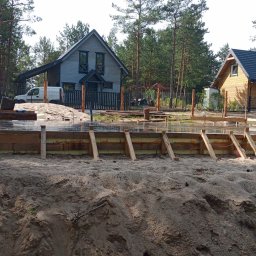 POL-HOME Marcin Polak - Wykop pod fundament z drewnianym szalunkiem i izolacją, w tle dwa domy jednorodzinne i las. Widoczny samochód osobowy i dostawczy.