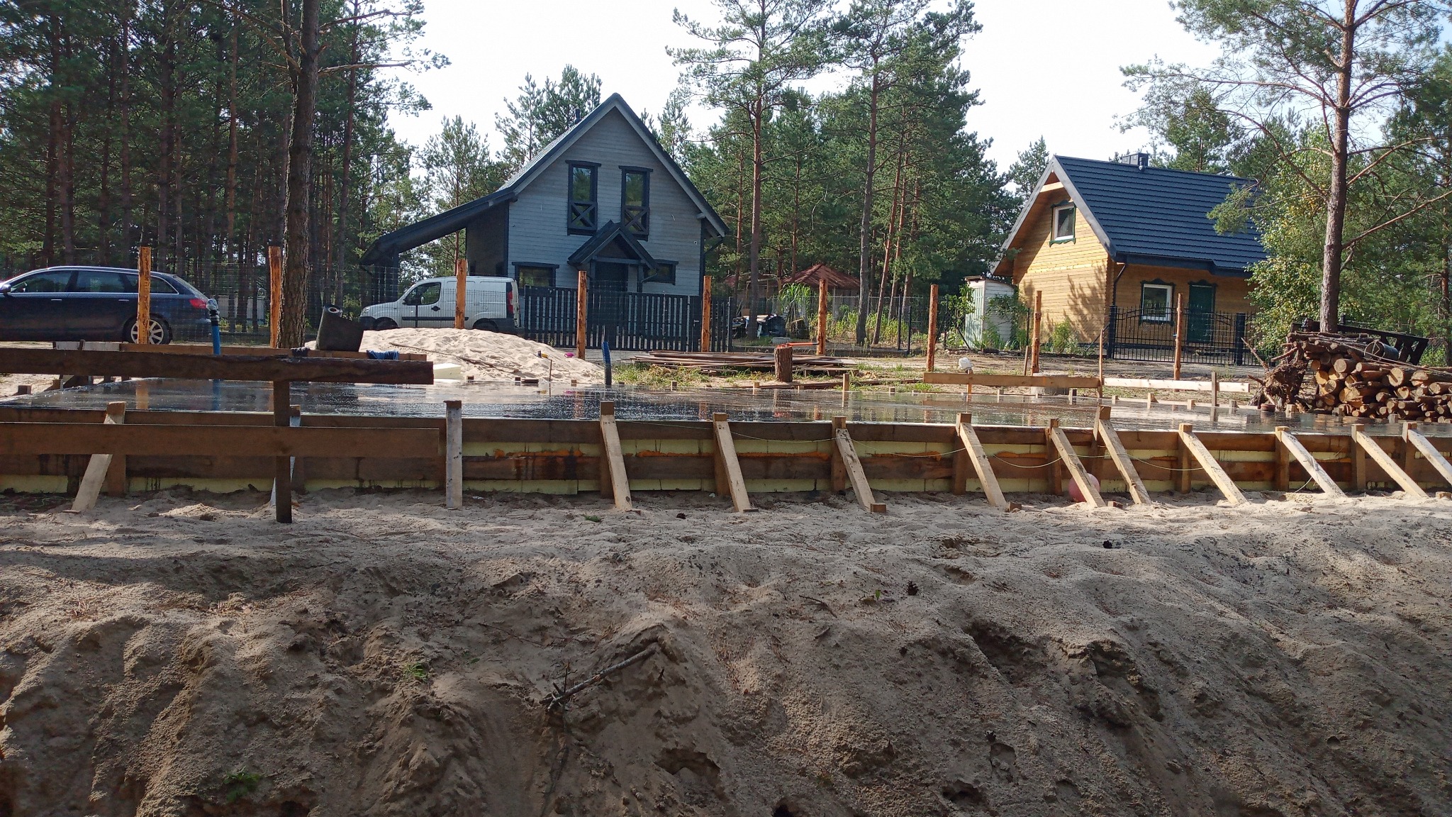 Wykop pod fundament z drewnianym szalunkiem i izolacją, w tle dwa domy jednorodzinne i las. Widoczny samochód osobowy i dostawczy.