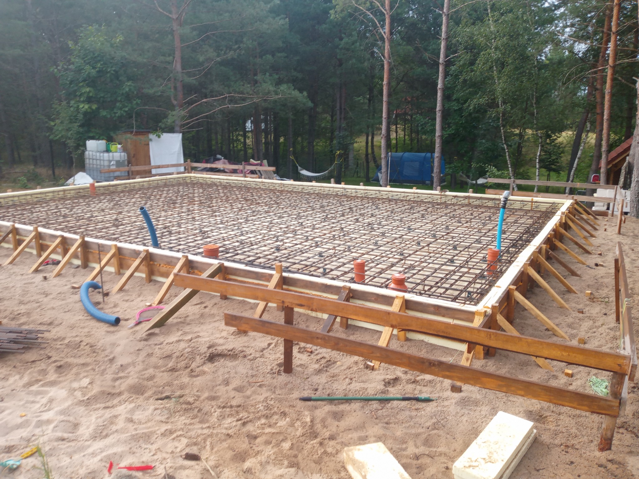 Zbrojenie fundamentu z drewnianym szalunkiem na piaszczystym podłożu, widok z góry. W tle drzewa i fragment zabudowy.