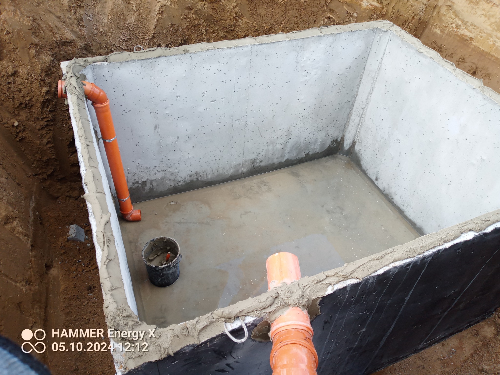 Betonowy zbiornik w ziemi z podłączonymi rurami kanalizacyjnymi i wiadrem, zabezpieczony hydroizolacją. Widok z góry na wykop.