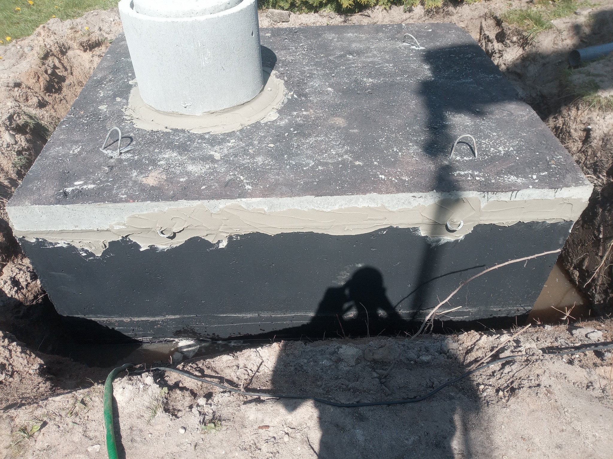 Betonowy zbiornik z włazem, uszczelniony i częściowo zakopany w ziemi. Widoczne haki montażowe i cień osoby robiącej zdjęcie.