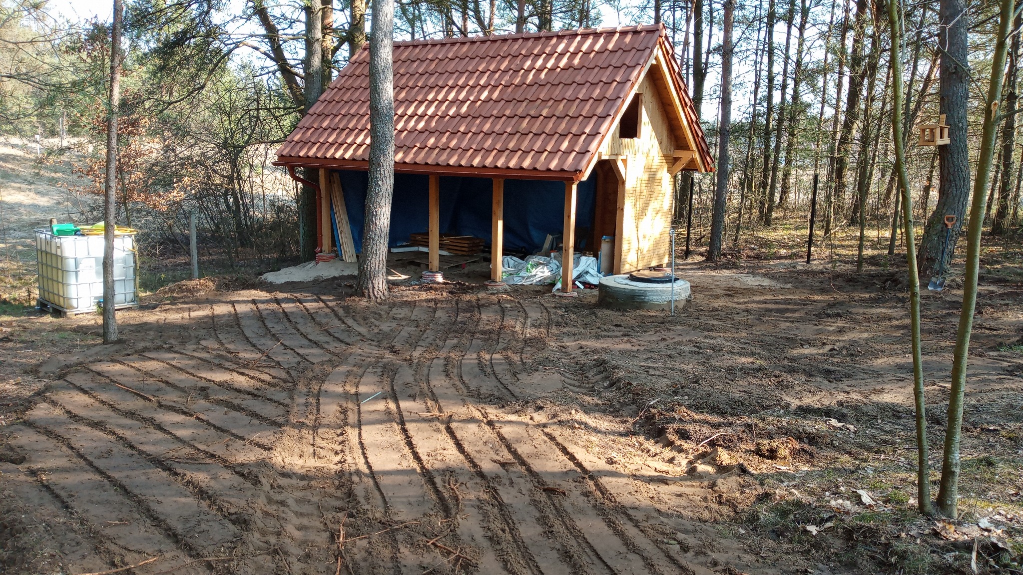 Drewniana altana z czerwoną dachówką w lesie, widoczne ślady po pracach ziemnych i studzienka kanalizacyjna. W tle drzewa i pojemnik na wodę.