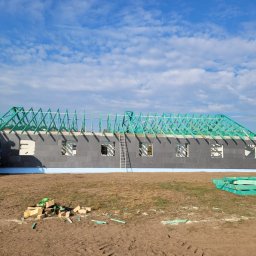 PJD CONSTRUCTION JAROSŁAW MICHAŁOWSKI - Wznoszenie domu szkieletowego: ściany z szarego bloczka i zielona więźba dachowa na tle błękitnego nieba z chmurami. Na pierwszym planie leżą materiały budowlane.