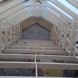 PJD CONSTRUCTION JAROSŁAW MICHAŁOWSKI - Wnętrze konstrukcji dachu z jasnego drewna, widok z dołu. Elementy połączone metalowymi płytkami. W tle mur z pustaków.