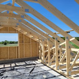 PJD CONSTRUCTION JAROSŁAW MICHAŁOWSKI - Wnętrze domu szkieletowego w budowie: widok na drewnianą konstrukcję ścian z oknem i więźbę dachową pod błękitnym niebem. Etap montażu.
