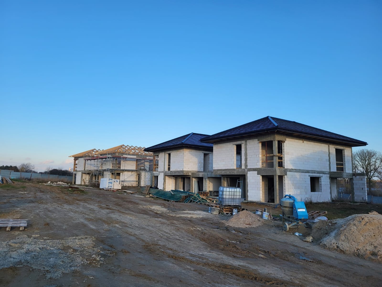 Budowa domów w stanie surowym otwartym: dwa budynki z dachem, trzeci w trakcie budowy więźby, białe ściany, błękitne niebo, ziemisty teren budowy.